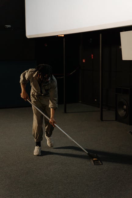 A professional cleaner from Carpet Cleaners Tooting is performing surface cleaning and sanitisation in an indoor auditorium or theater setting, using a vacuum cleaner with a yellow and black body. The