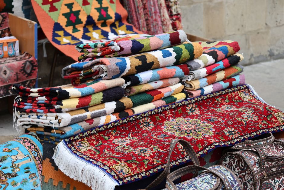 A display of various woven rugs and carpets with intricate patterns, hanging on a wall and laid out on the floor within a shop environment. The rugs feature diverse geometric and traditional motifs, w